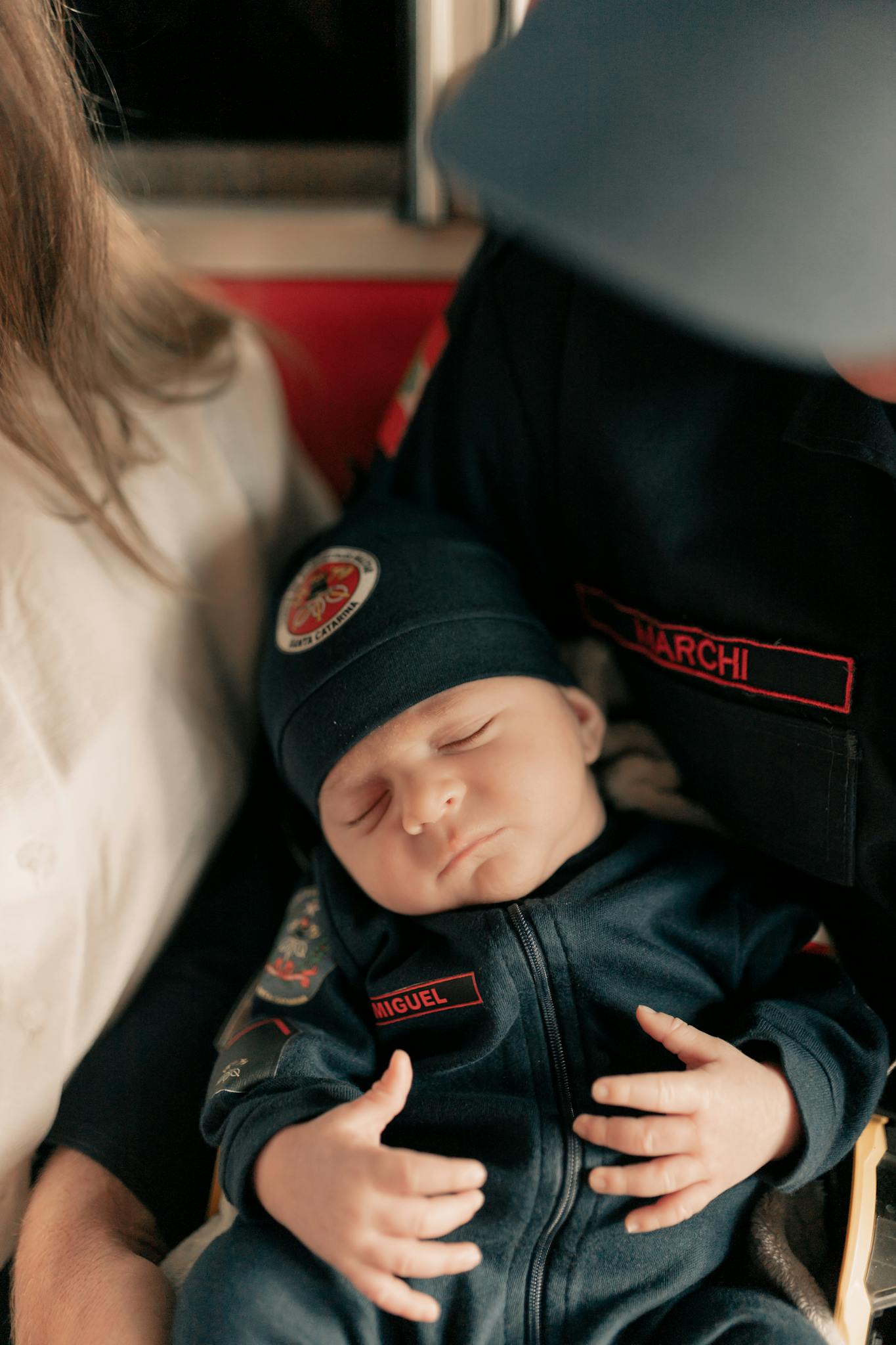 Adorable baby asleep in firefighter attire, embraced by parents.