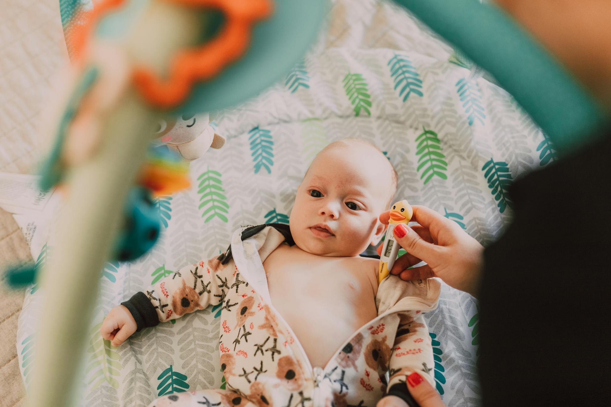 Cute baby lying indoors with a thermometer, wearing a patterned onesie.