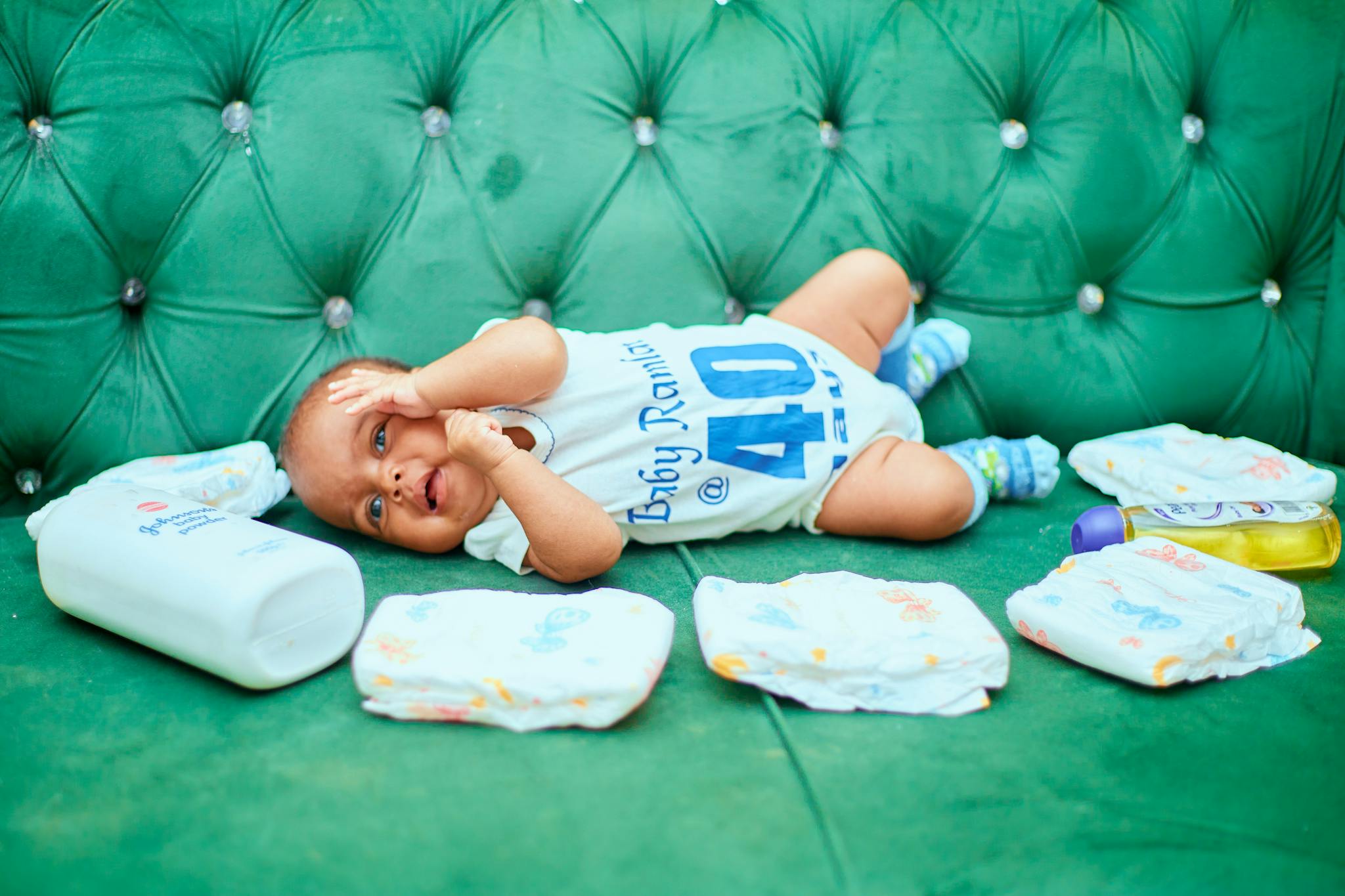 Cute baby lying on a green sofa surrounded by diapers and baby products.